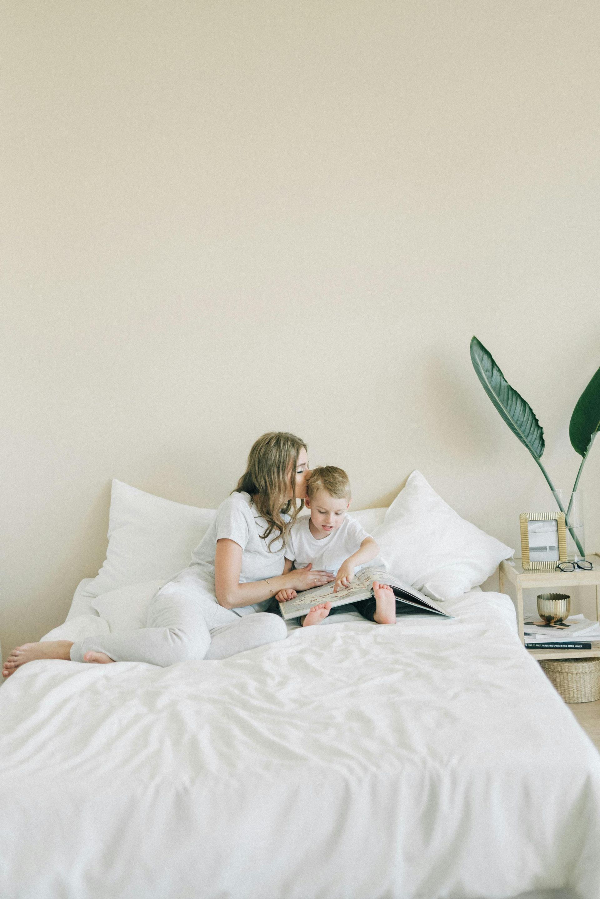 Une femme et un enfant lisent un livre sur un lit dans une pièce lumineuse.