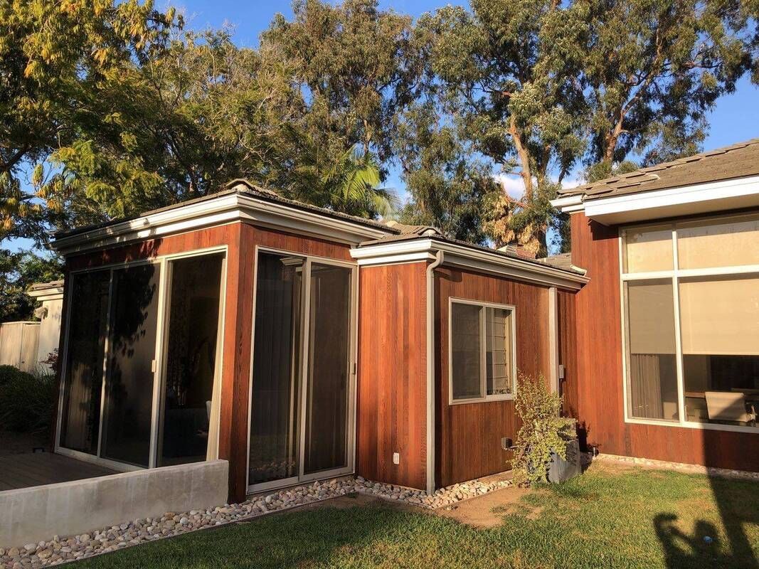A sunroom addition with reddish-brown siding adjoins a house; a small window and screen doors are visible.
