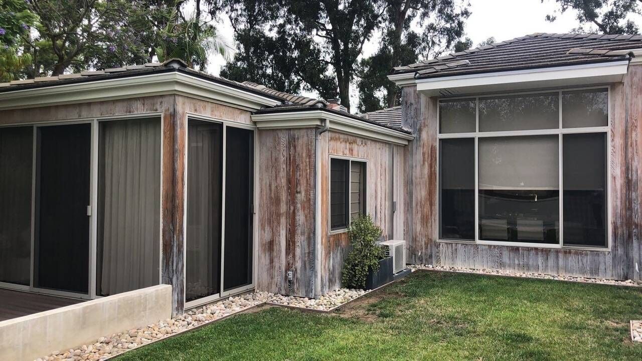 Exterior of a house with weathered stucco siding, tile roof, large windows, and a small patch of grass.