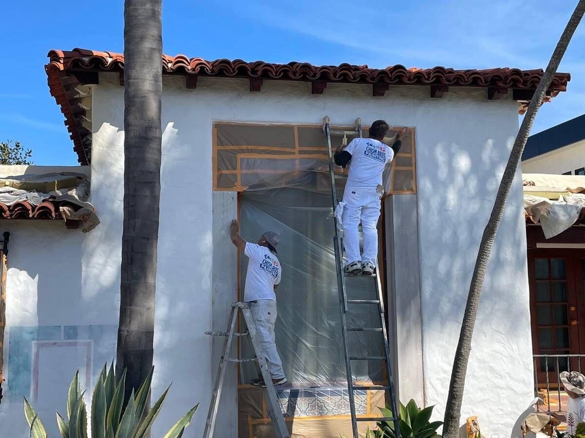 Two painters in white coveralls, using ladders, painting a stucco building.