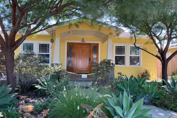 Yellow house with brown door, flanked by windows. Landscaping in front includes succulents and trees.