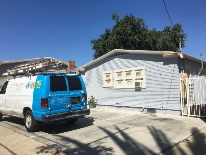 Blue van parked in front of a light blue house. Ladder on van roof. Clear sky.