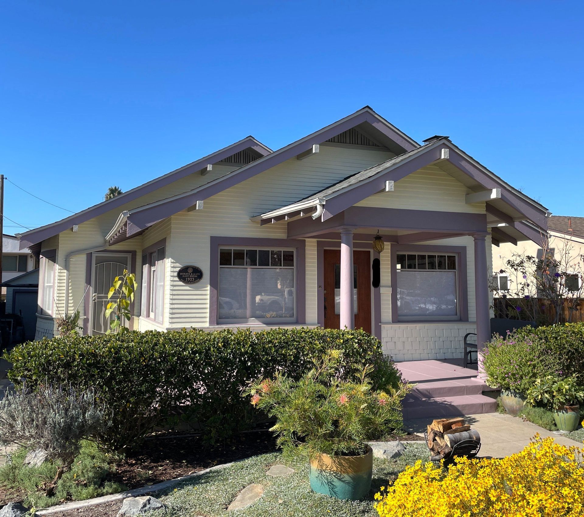 Cottage-style house with light yellow siding and lavender trim, surrounded by landscaping, under a blue sky.