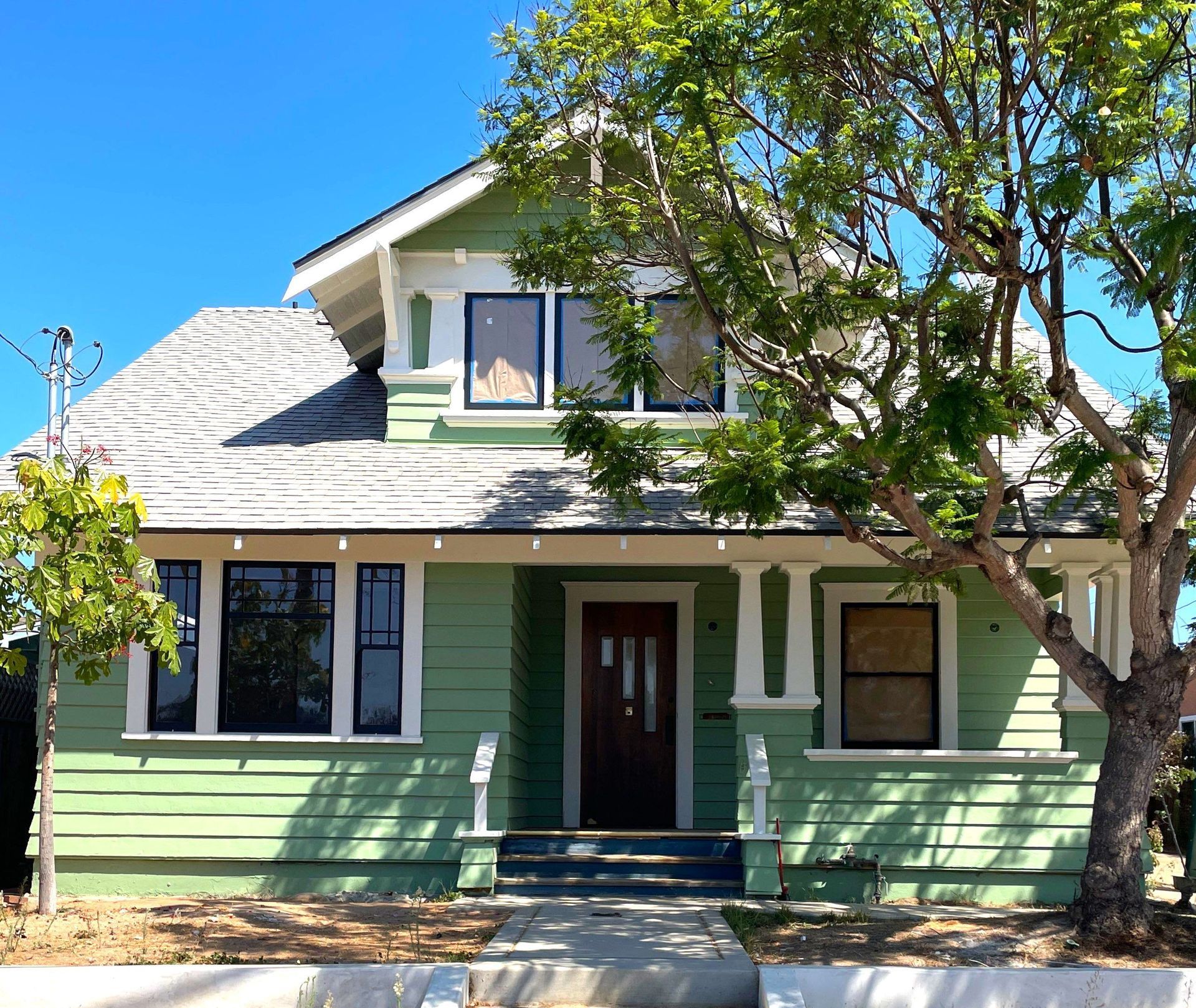 Green Craftsman house with white trim, dark windows, and a brown door; a tree shades the front.