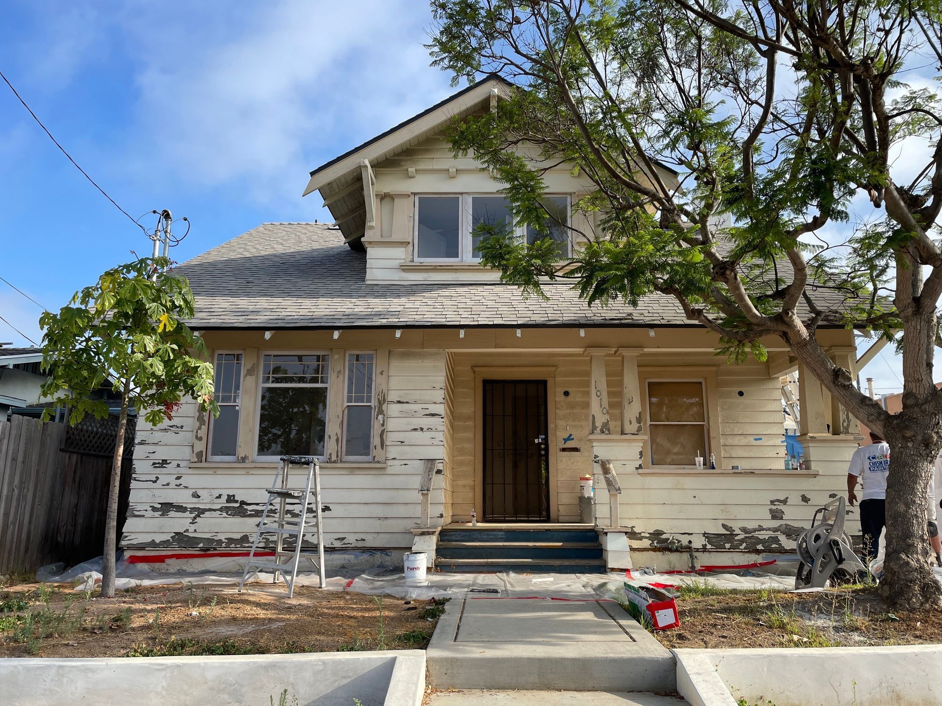 Old, off-white house with peeling paint, front door, and gray roof. Trees in front, cloudy sky. Person on the right.