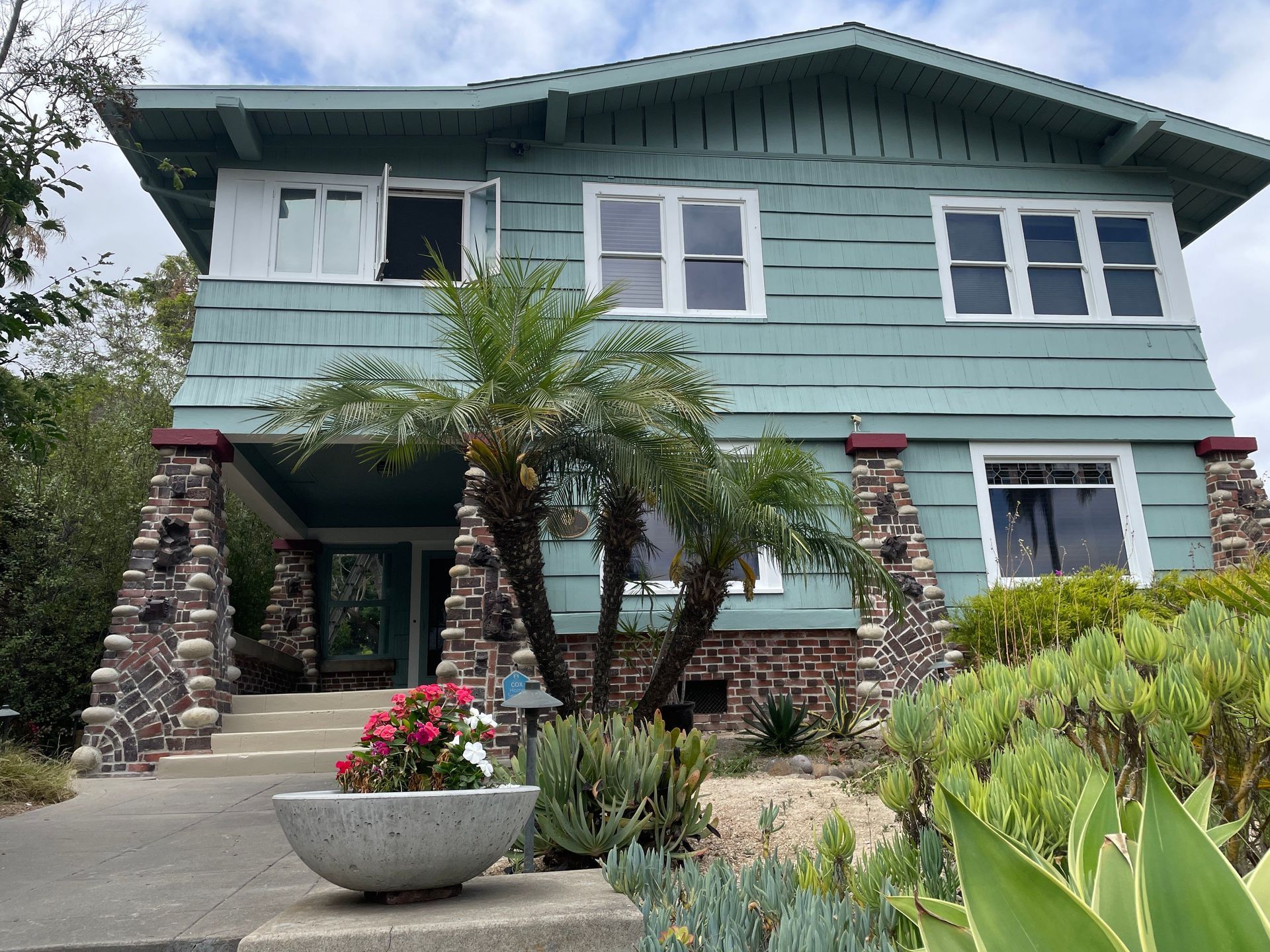 Two-story teal house with stone columns, palm trees, and a concrete planter.
