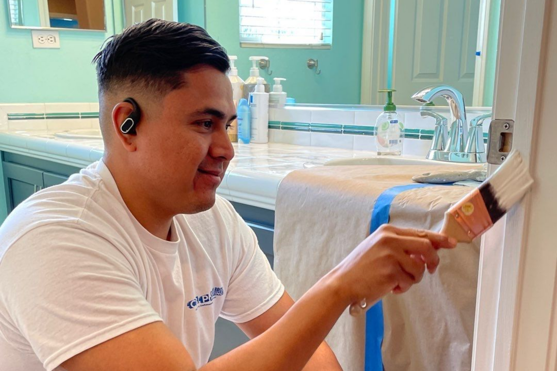 Man in white shirt painting a bathroom door frame with a brush. He is smiling while listening to earbuds.