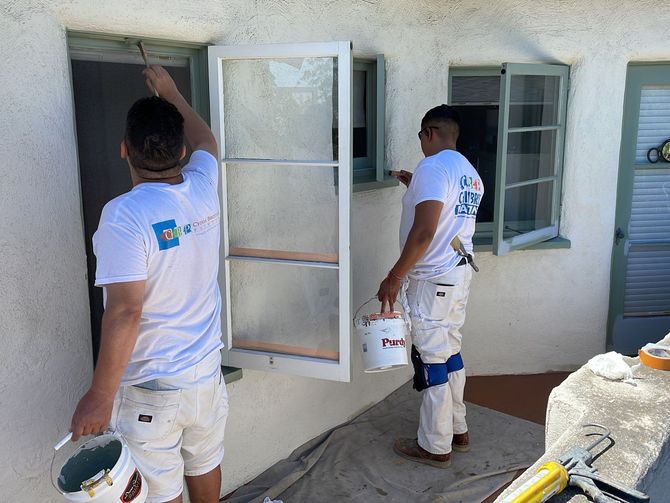 Two painters in white overalls paint window frames on a stucco building.