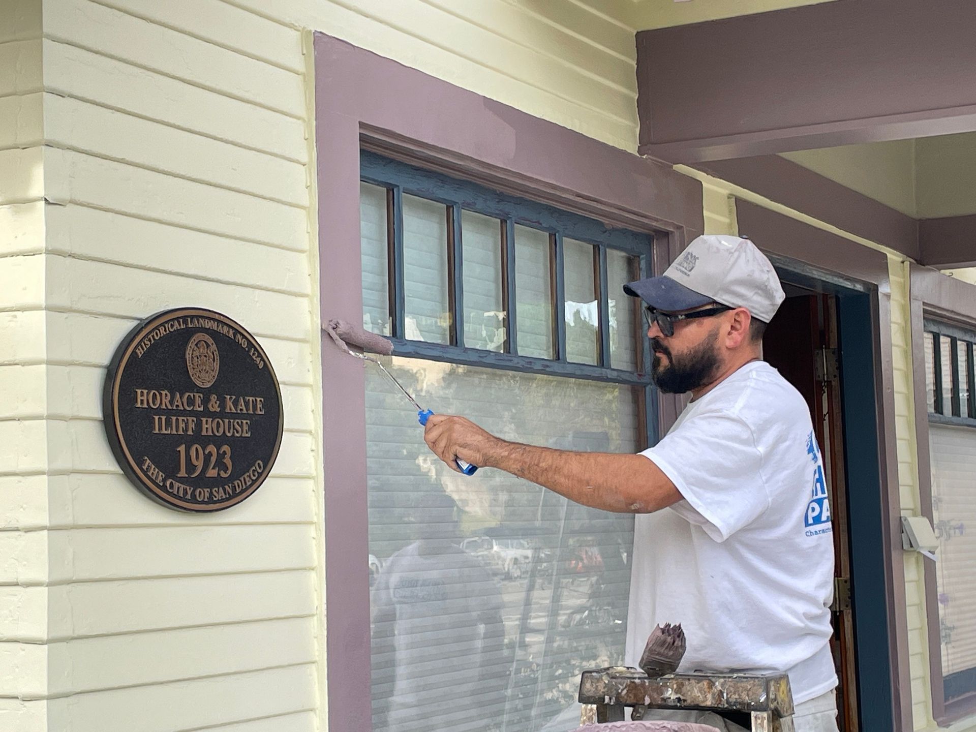 Man painting window trim on a light-colored house. Dark bronze plaque nearby.