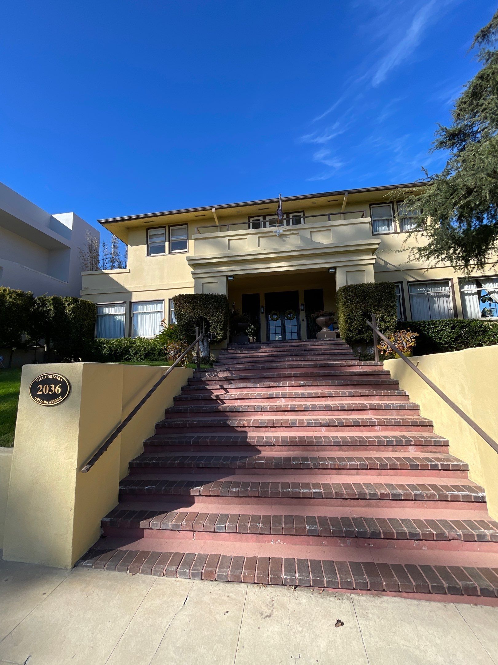 Yellow building with stairs leading to the entrance, flanked by bushes and a blue sky.