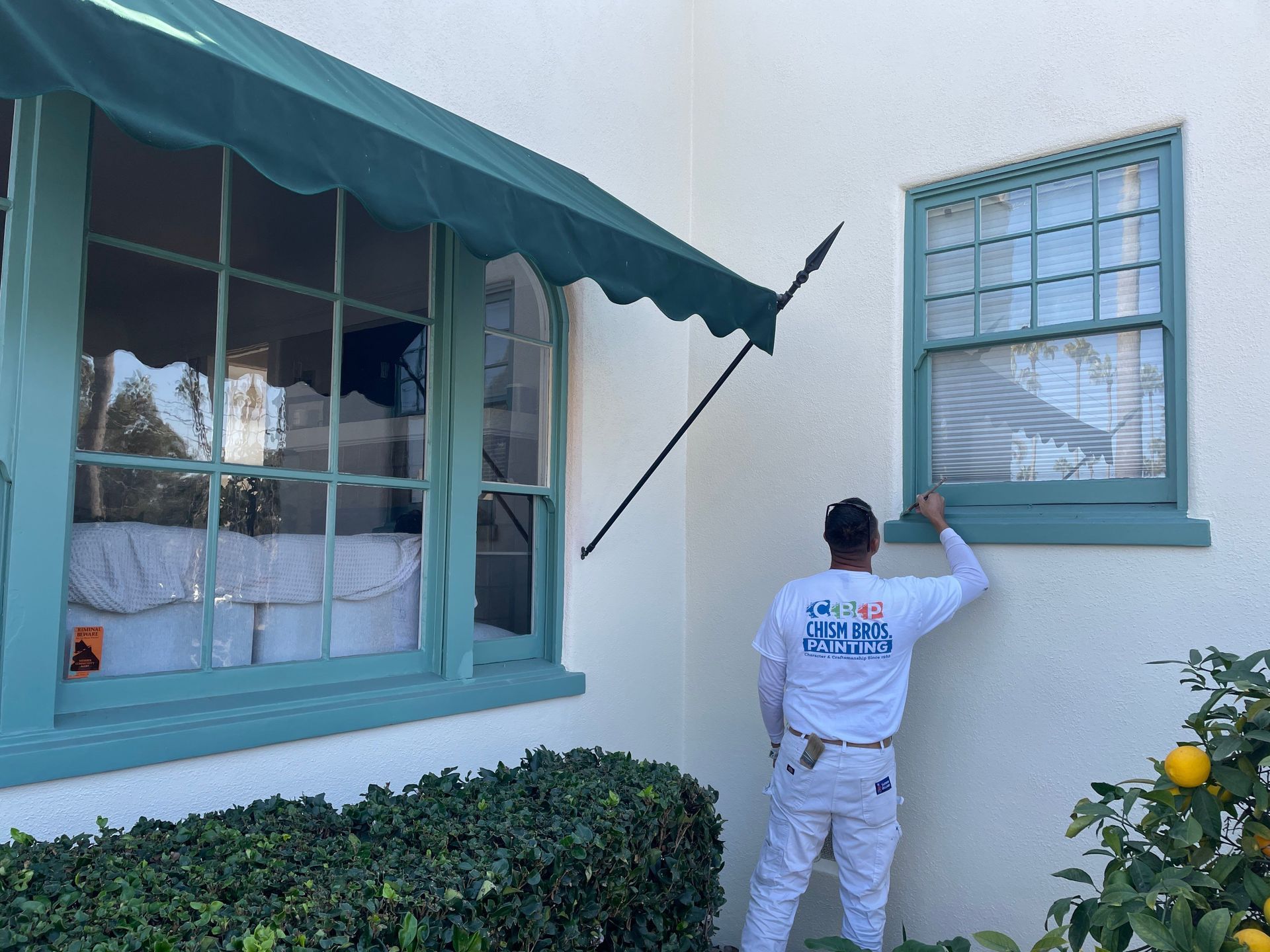 A person painting window trim on a white house, green awning and trim, sunny day.