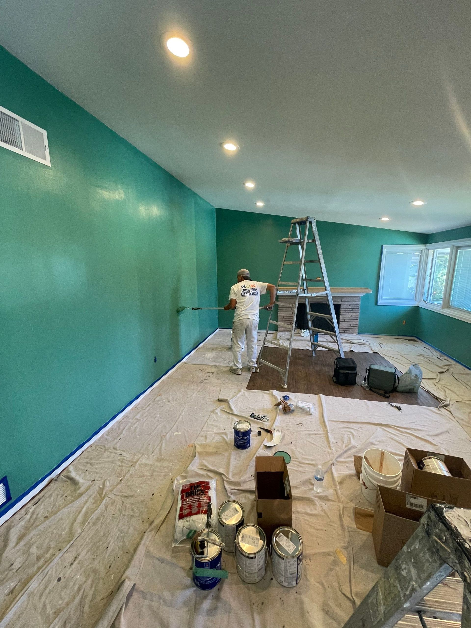 A person paints a room teal. Paint cans and drop cloths are on the floor.