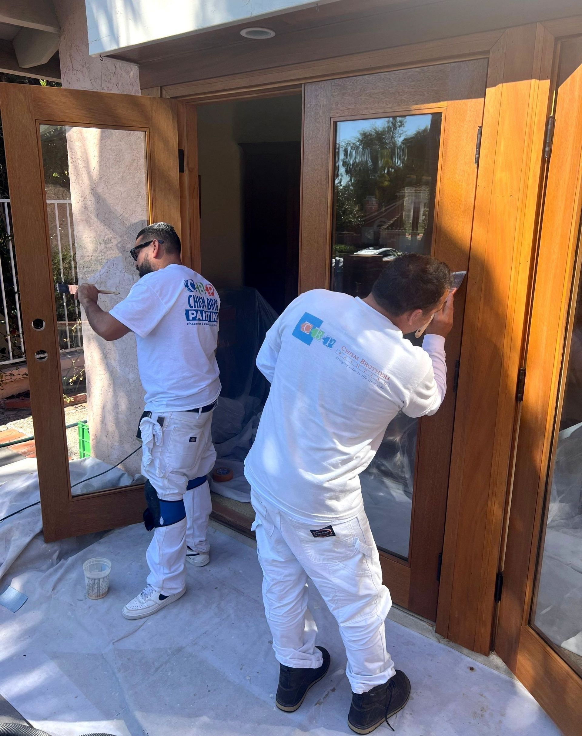 Two painters in white coveralls, working on wooden double doors outdoors.