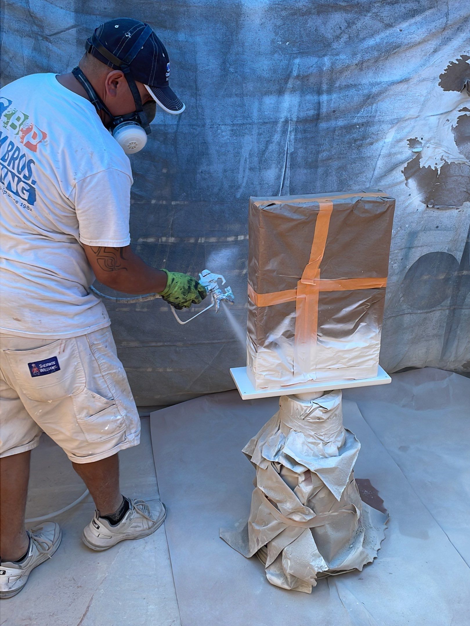 A person wearing a mask sprays paint on a brown, taped box on a pedestal in an enclosed space.
