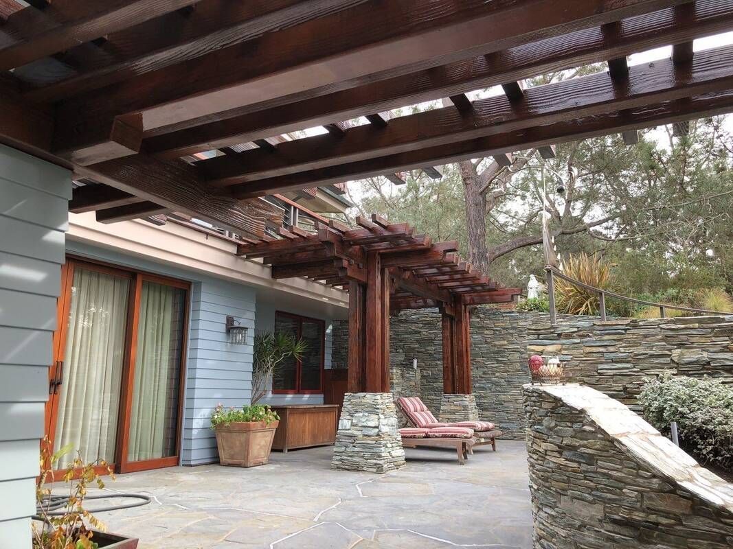 Wooden pergola over a stone patio with outdoor furniture, next to a house with sliding glass doors.