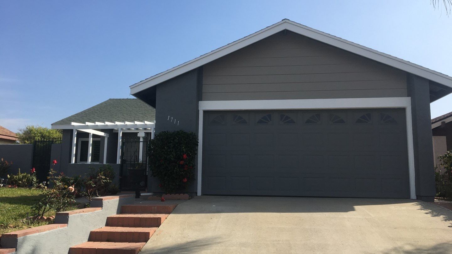 Gray house with a two-car garage, red brick steps, and a pergola with green roof.