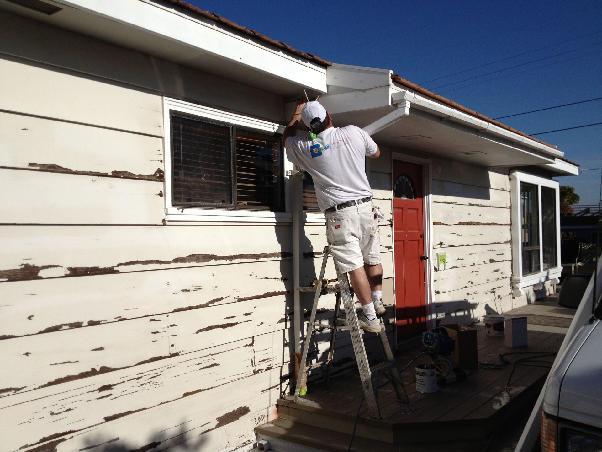 Person on ladder installing gutter on a white house with peeling paint.