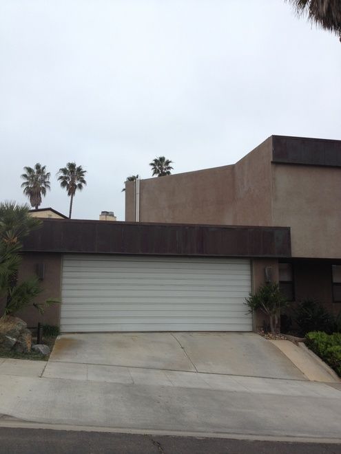 Beige house with a white garage door, palm trees, and a concrete driveway under a cloudy sky.