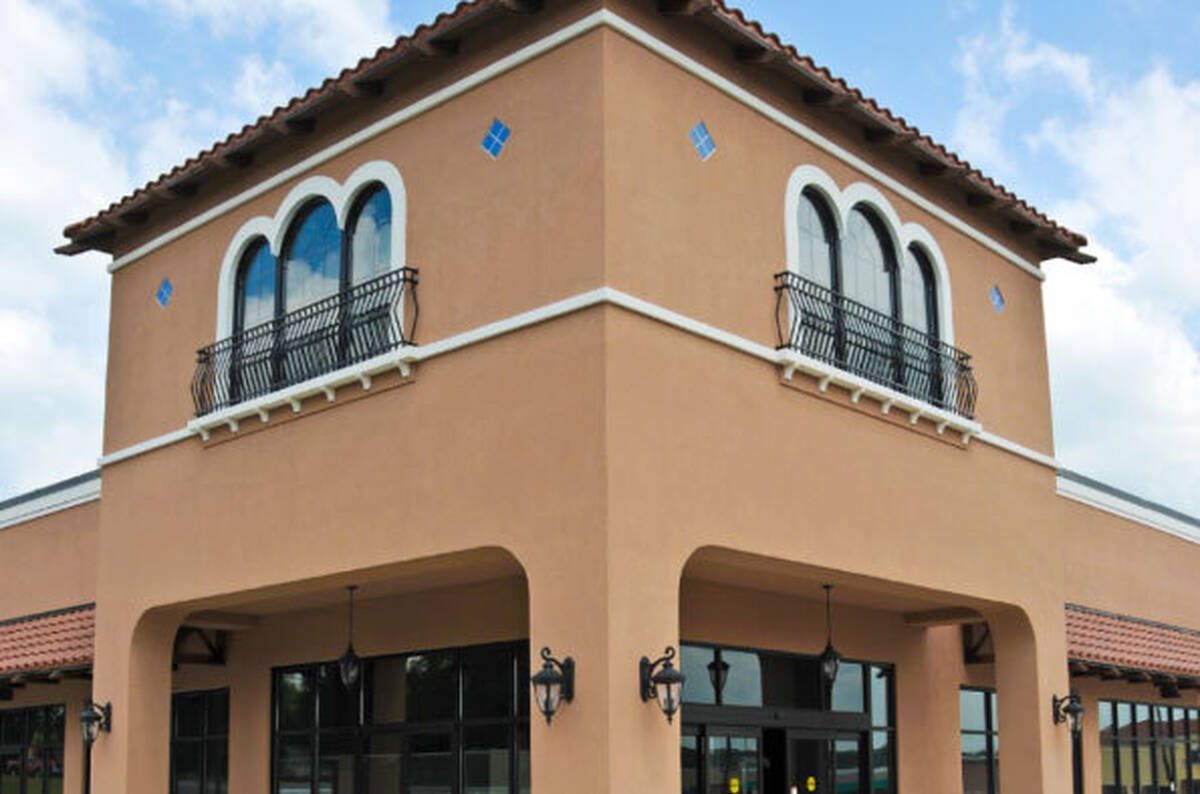 Tan stucco building with arched windows, black iron balconies, and tiled roof against a blue sky.
