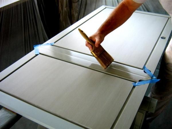 Person painting a white door with a paintbrush, blue tape, and a light-colored surface.
