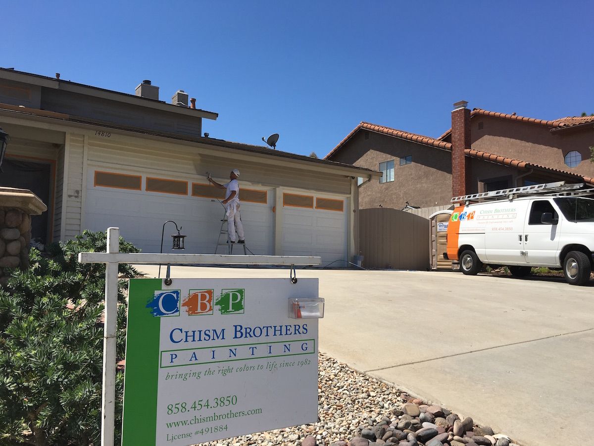 Painter on a ladder painting a beige house; a white service van parked in the driveway.