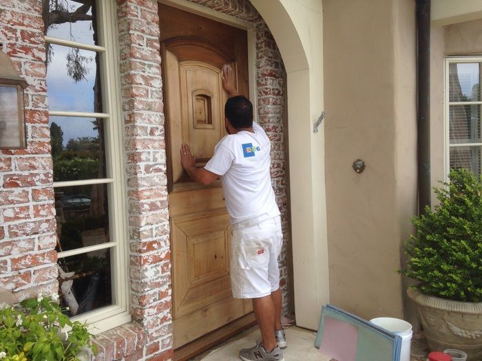 Man in white clothes inspecting a wooden door; brick and beige exterior.