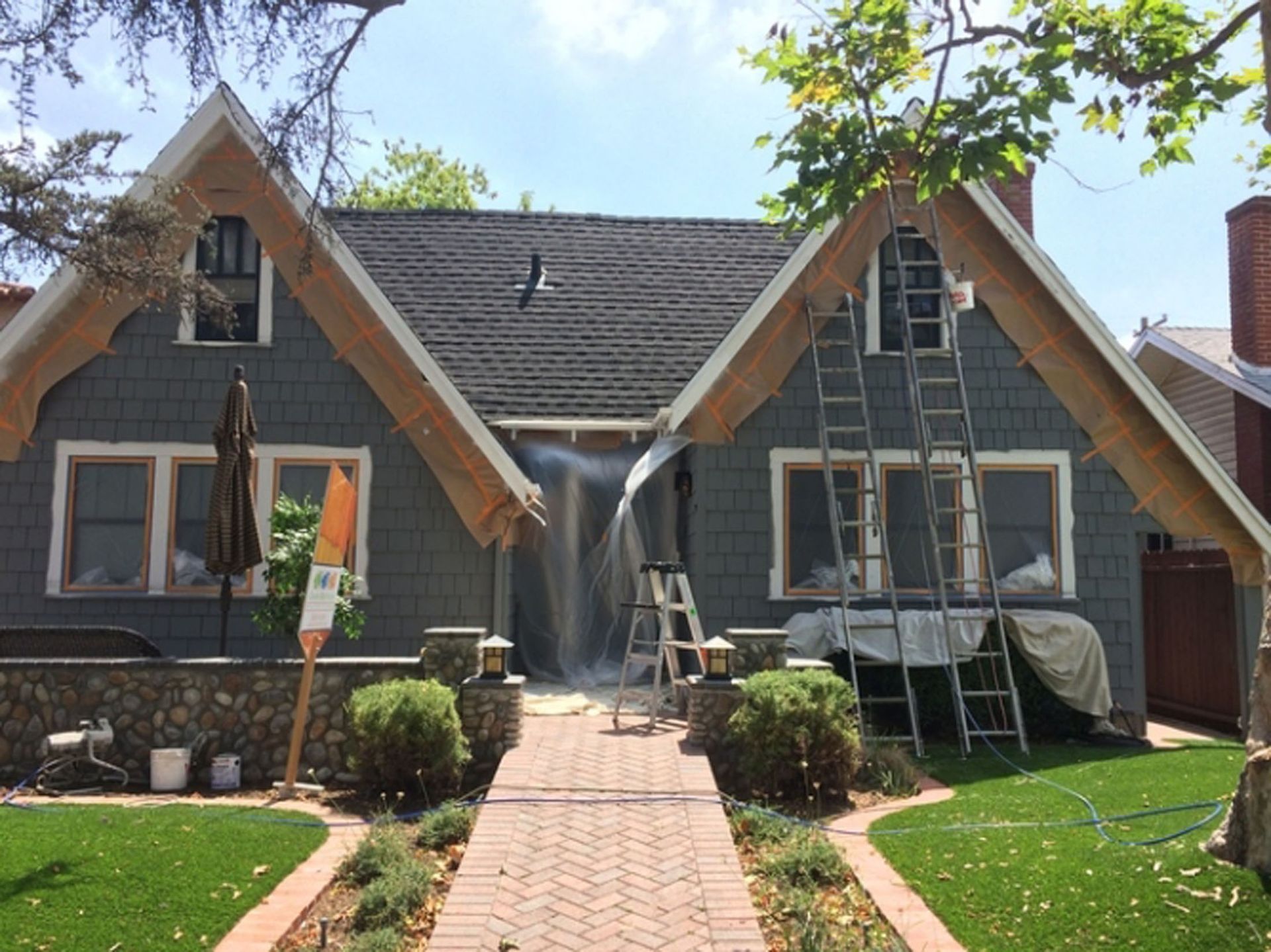 House exterior being painted, with ladders, plastic sheeting, and brick walkway. Gray siding, tan trim.