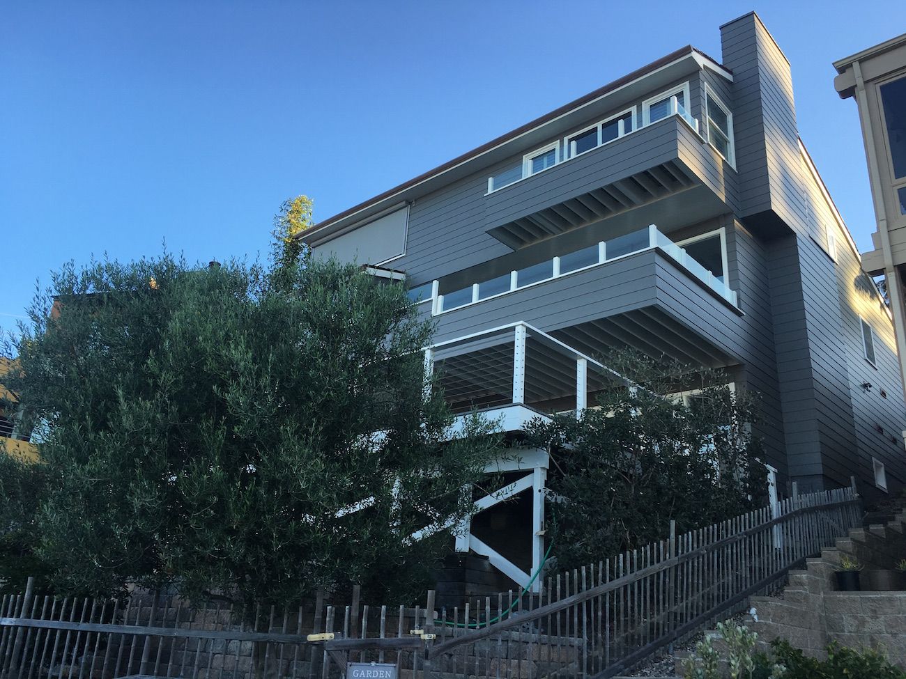 Three-story gray house with decks and white railing, nestled on a hillside with a wooden fence.
