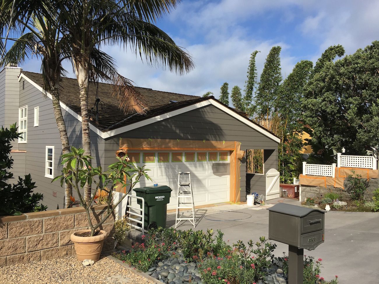Garage under construction on a gray house with a palm tree, a green trash bin, and a mailbox in the driveway.