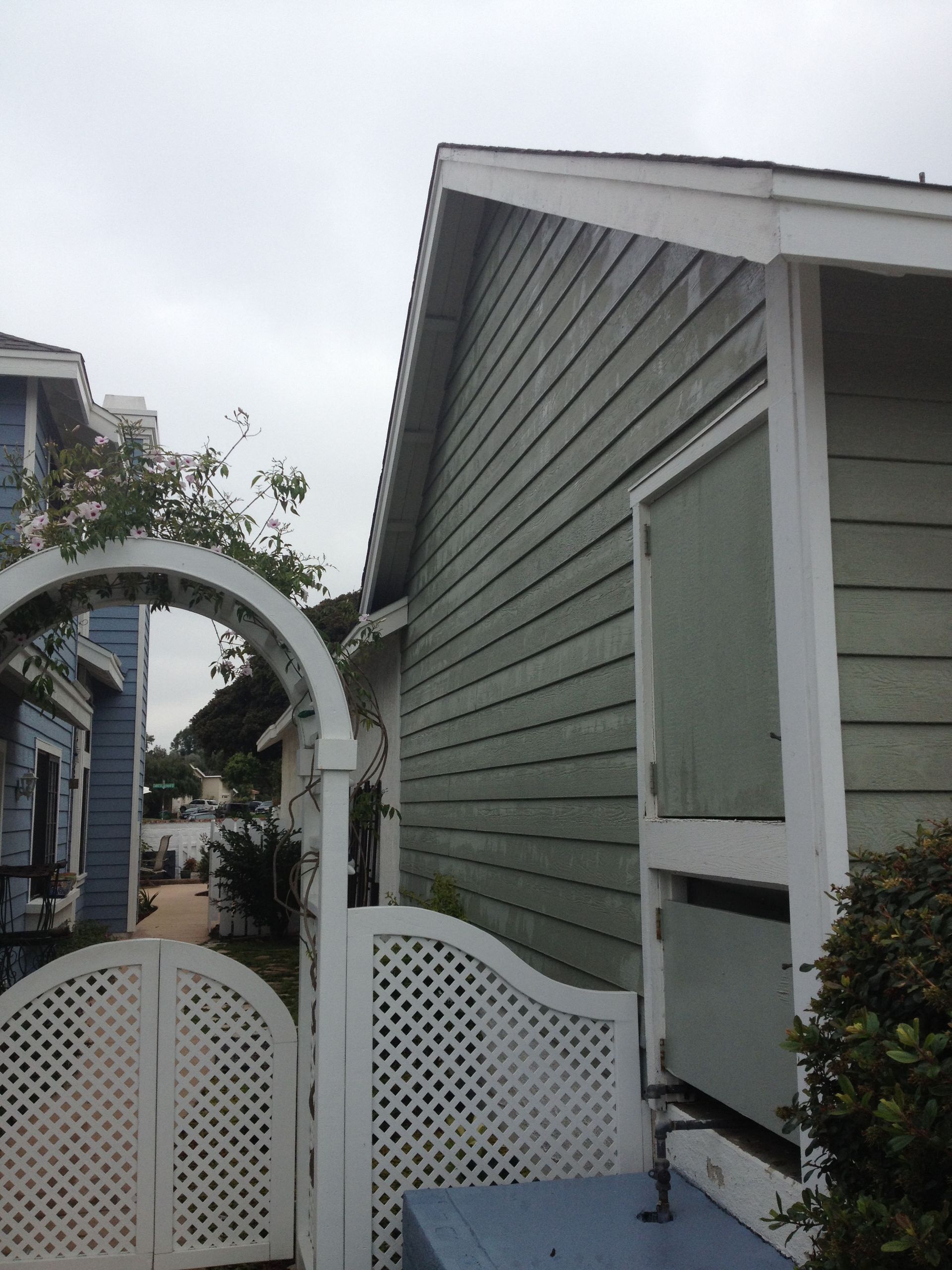 Green house siding next to a white arched gate, blue house in the distance, overcast sky.