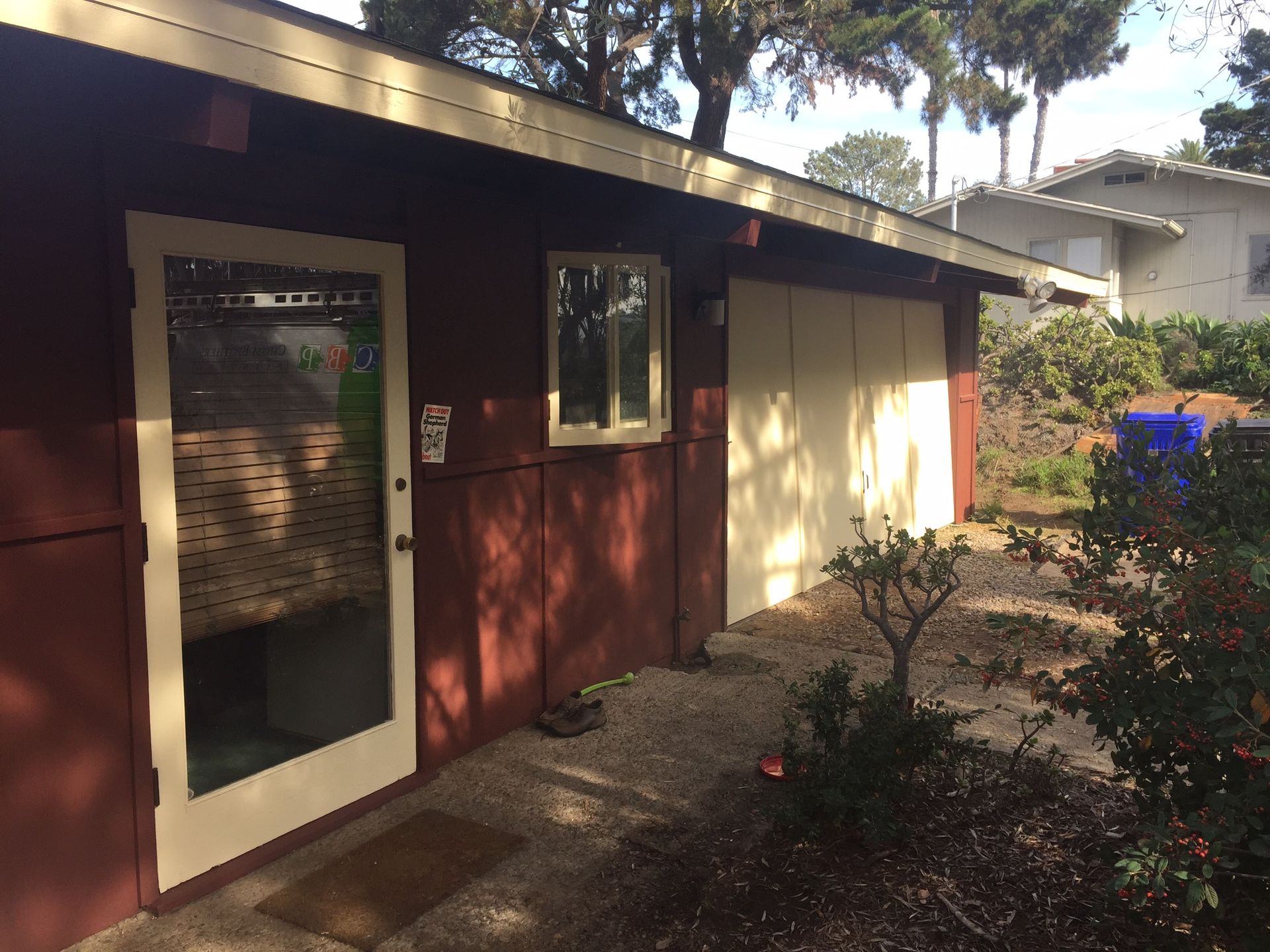 Red building with a glass door, window, and closed garage door; beige trim; gravel path, bushes, and trees.