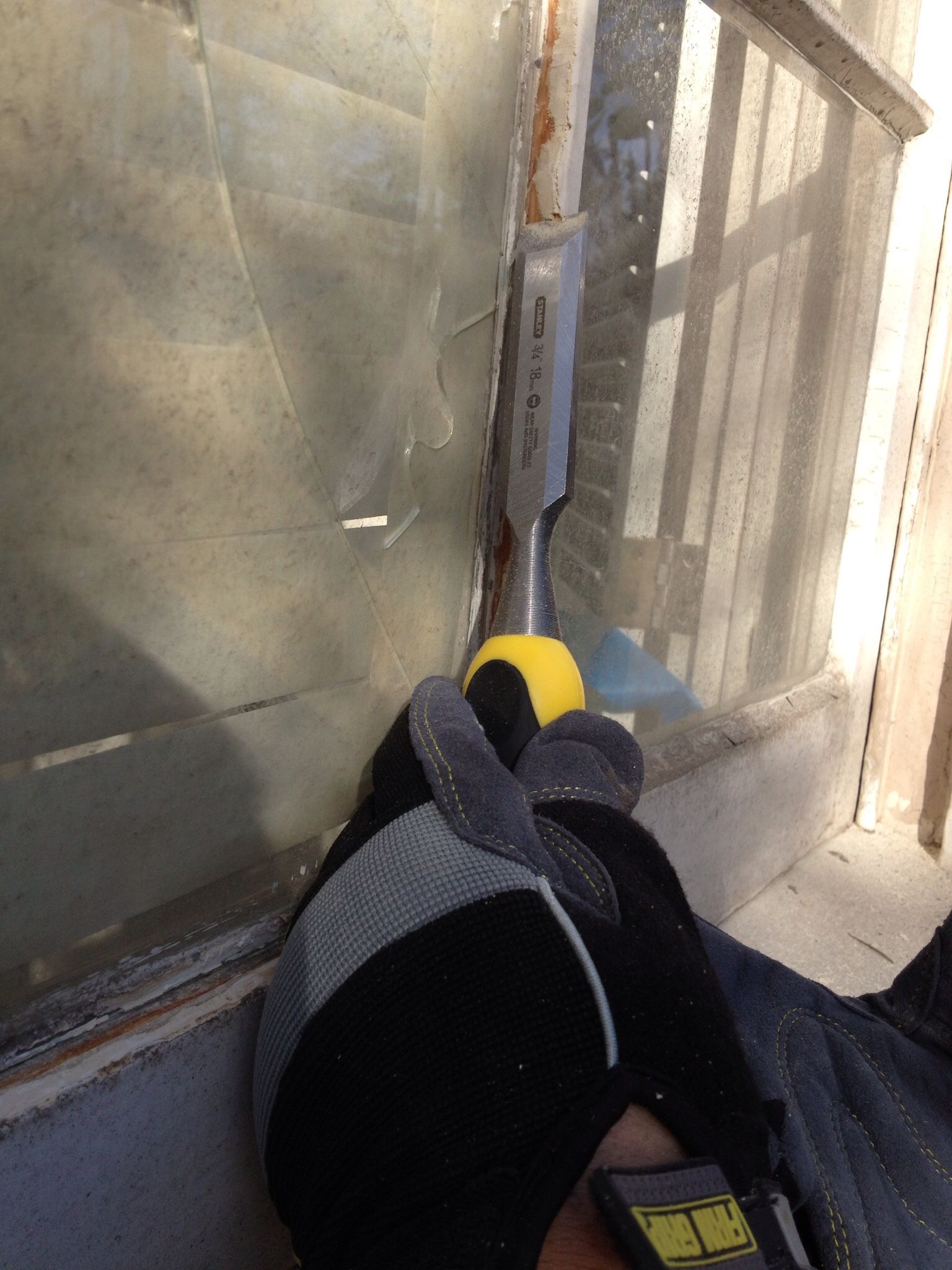 A gloved hand uses a chisel to remove something from a window frame.