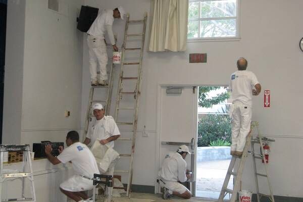 Five people in white overalls painting a room, using ladders and various tools.