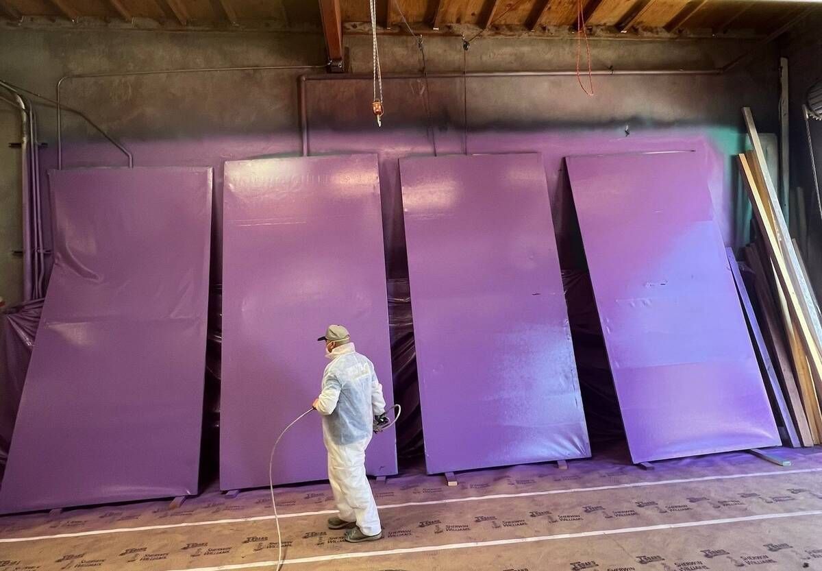 A person in protective gear sprays purple paint onto large panels in a workshop.