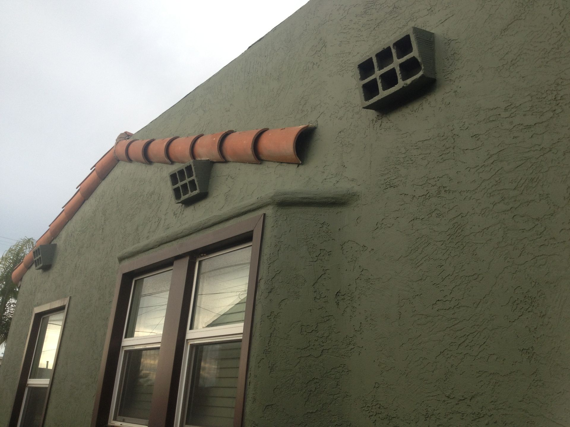 Green stucco house with terracotta roof tiles, brown window trim, and decorative block features.