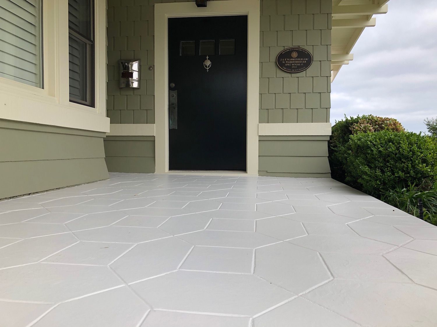 Gray painted porch with patterned design leading to a black door with green siding and a bush.