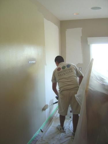 Person painting an interior wall, using a paint sprayer. White walls, painter in tan clothes.