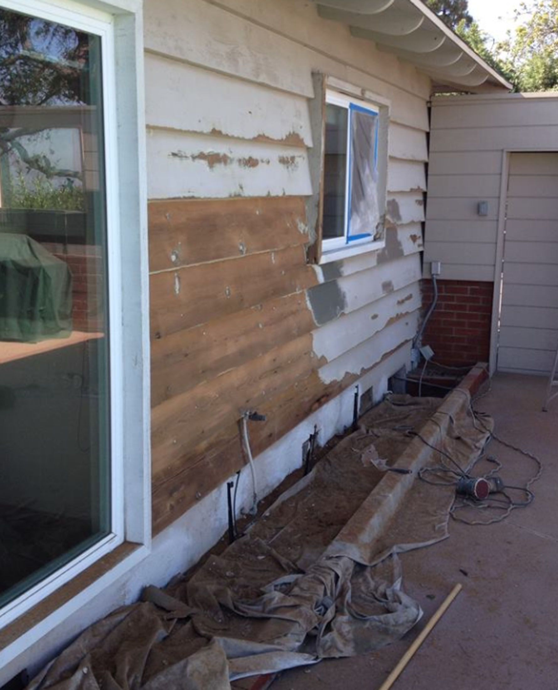 Exterior house wall, partially stripped of siding, showing wood, a window, and tarp on the ground.