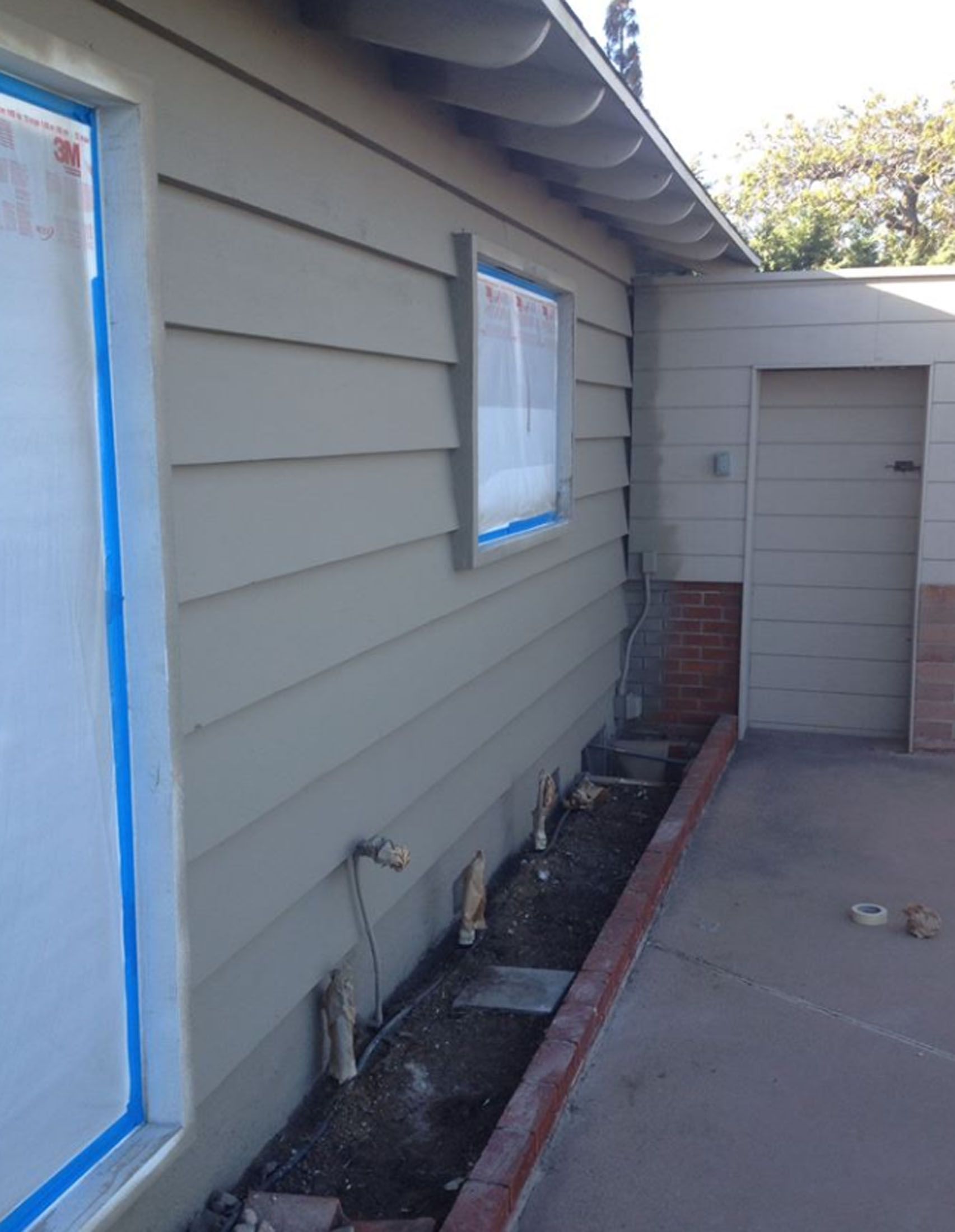 Exterior wall with painted siding, boarded window, and brick edging along the base; next to a concrete patio.