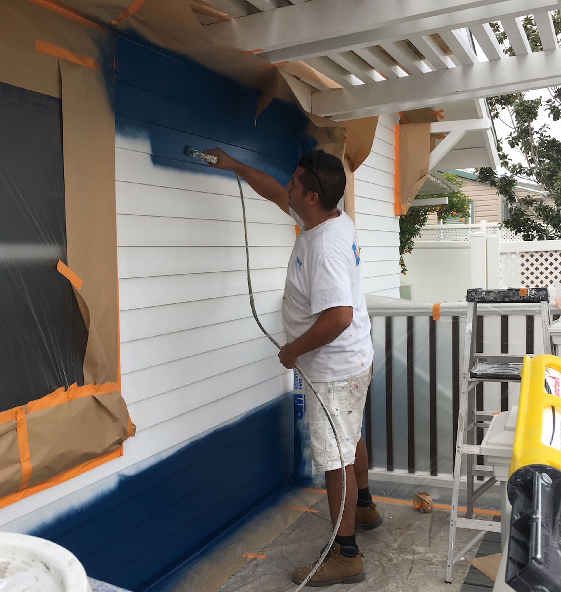 Man painting a white exterior wall blue with a spray gun, using masking paper.