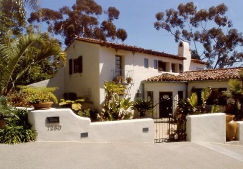 White stucco home with red tile roof, surrounded by greenery and a low wall.