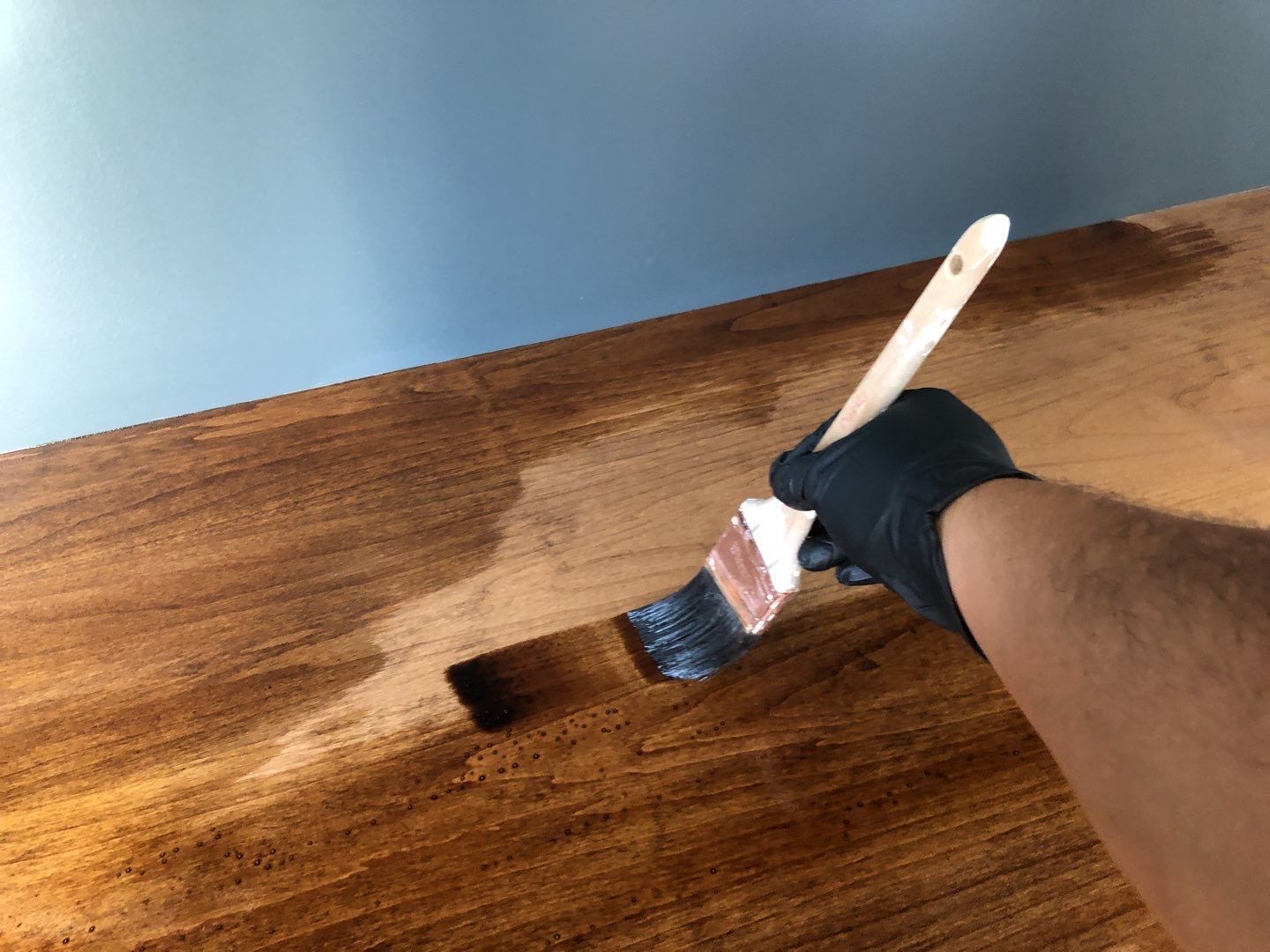 Hand wearing black glove staining wooden surface with a brush; blue wall in background.