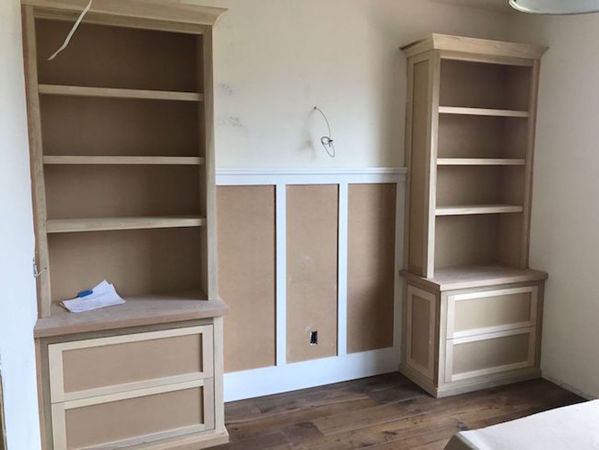 Two unfinished wooden bookshelves, one on either side of a partially paneled wall, on a wood floor.