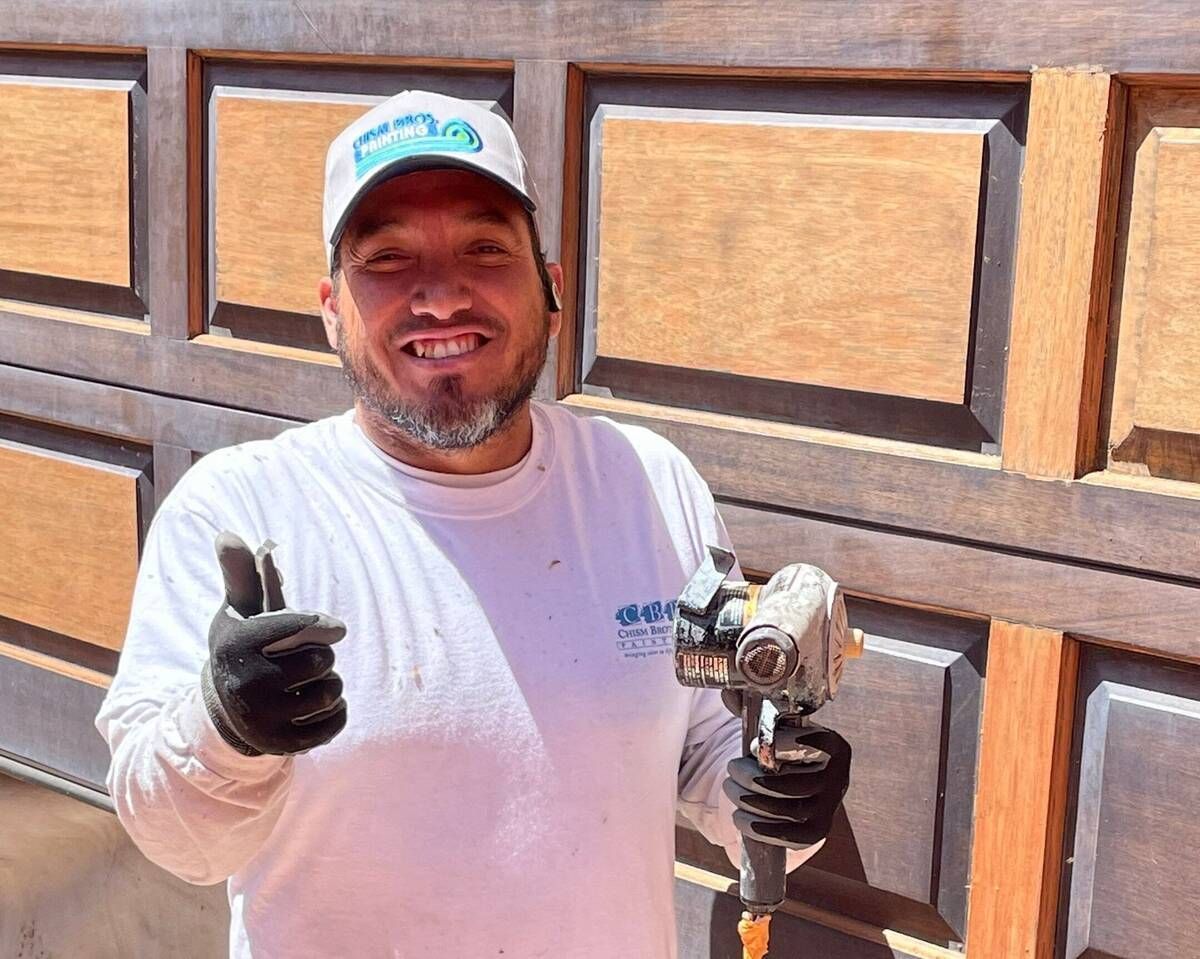 Man in hat and gloves gives thumbs up, holding tool in front of wooden door.