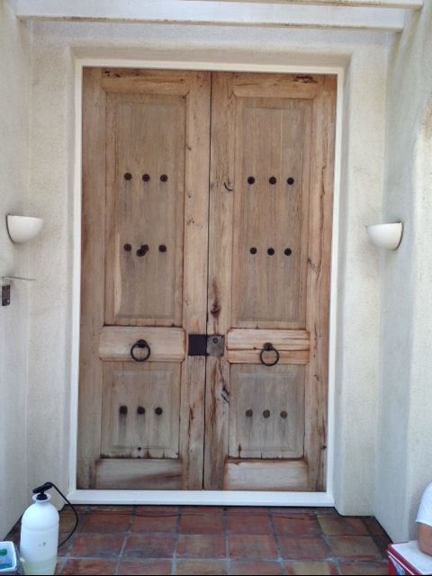 Wooden double doors with metal studs and ring handles, surrounded by white trim and sconces on a tan wall.