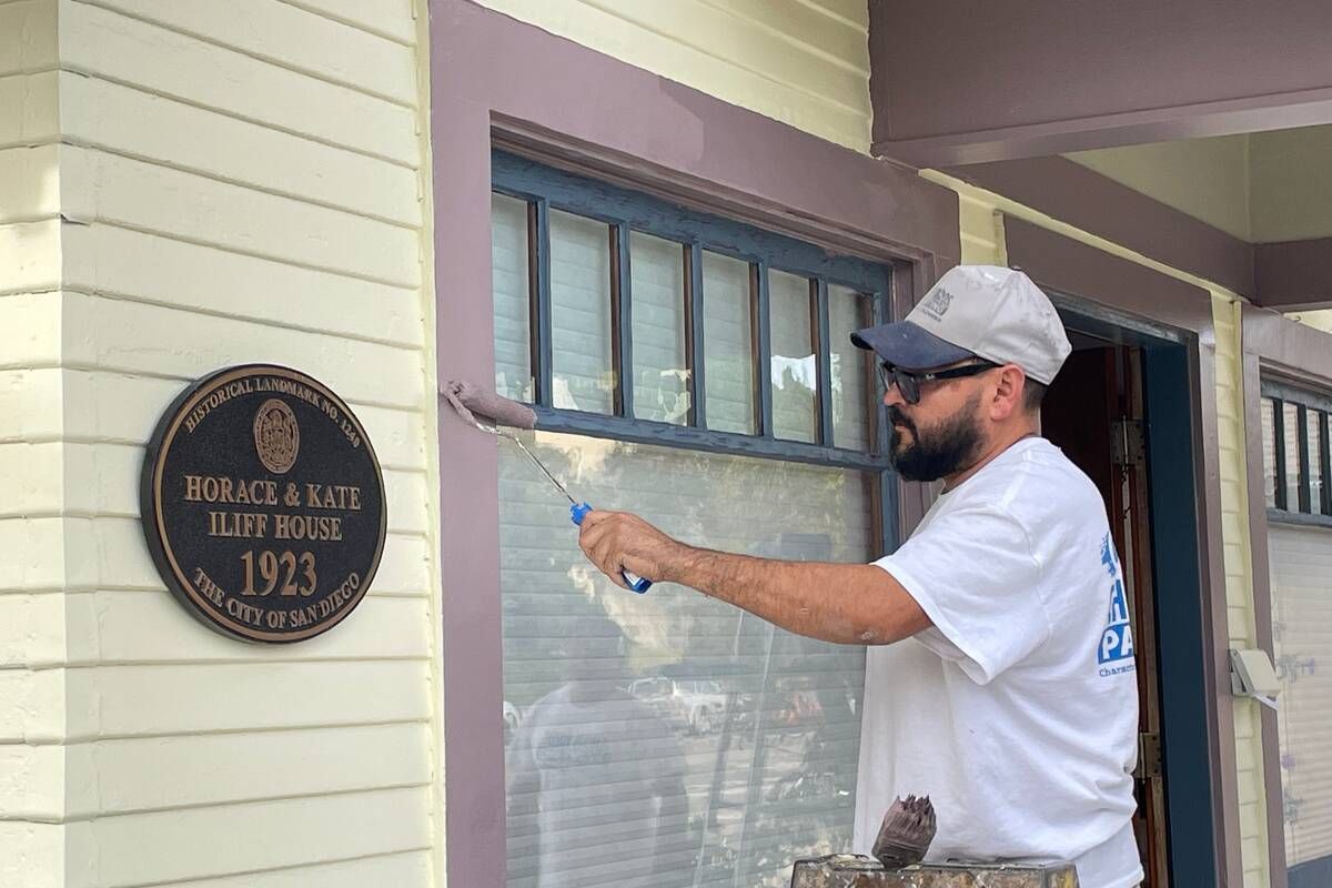 Man paints trim around a window on a house with a historic plaque.