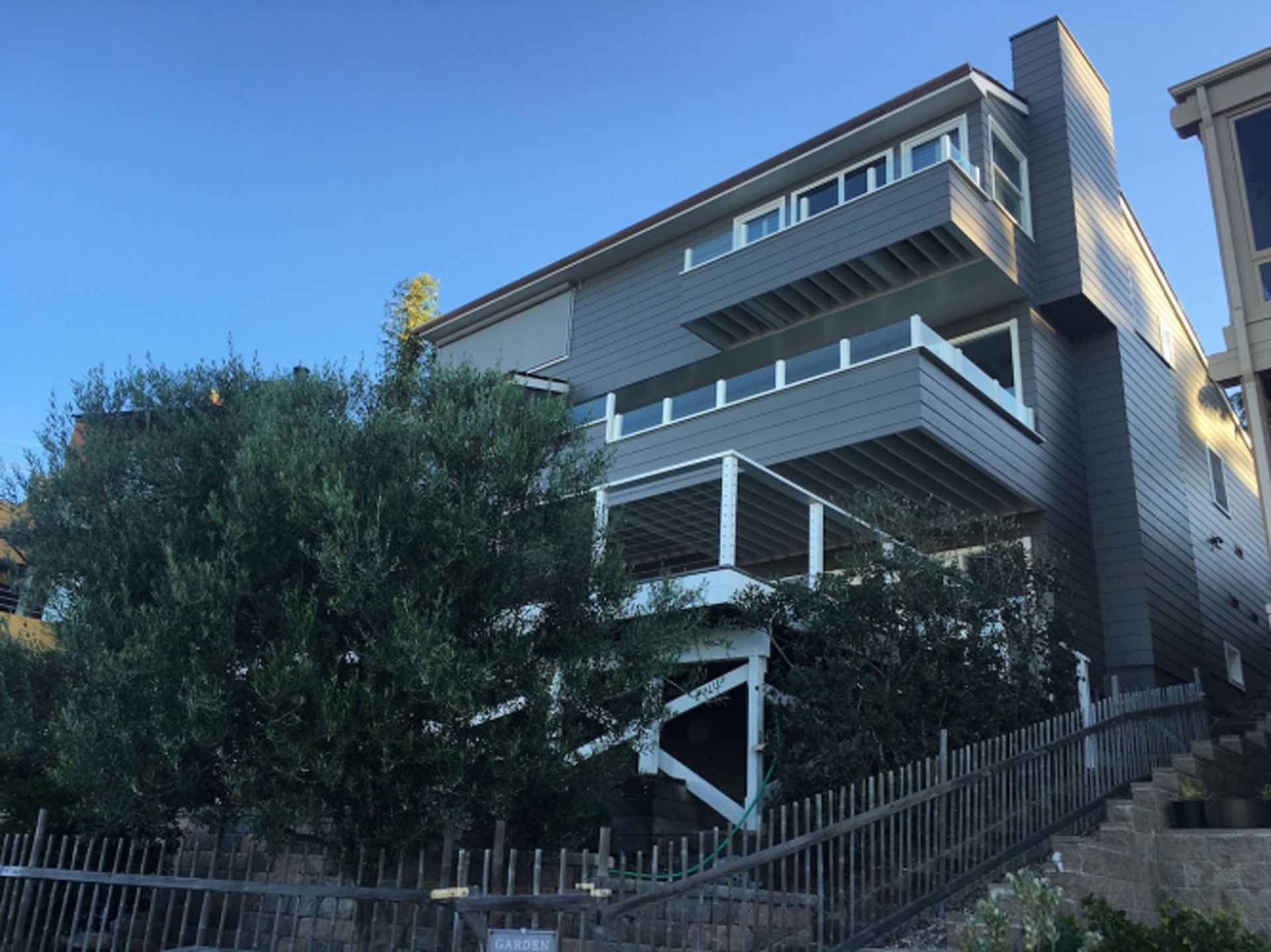 Multi-level house with gray siding and multiple decks, set on a hillside behind a fence and greenery.