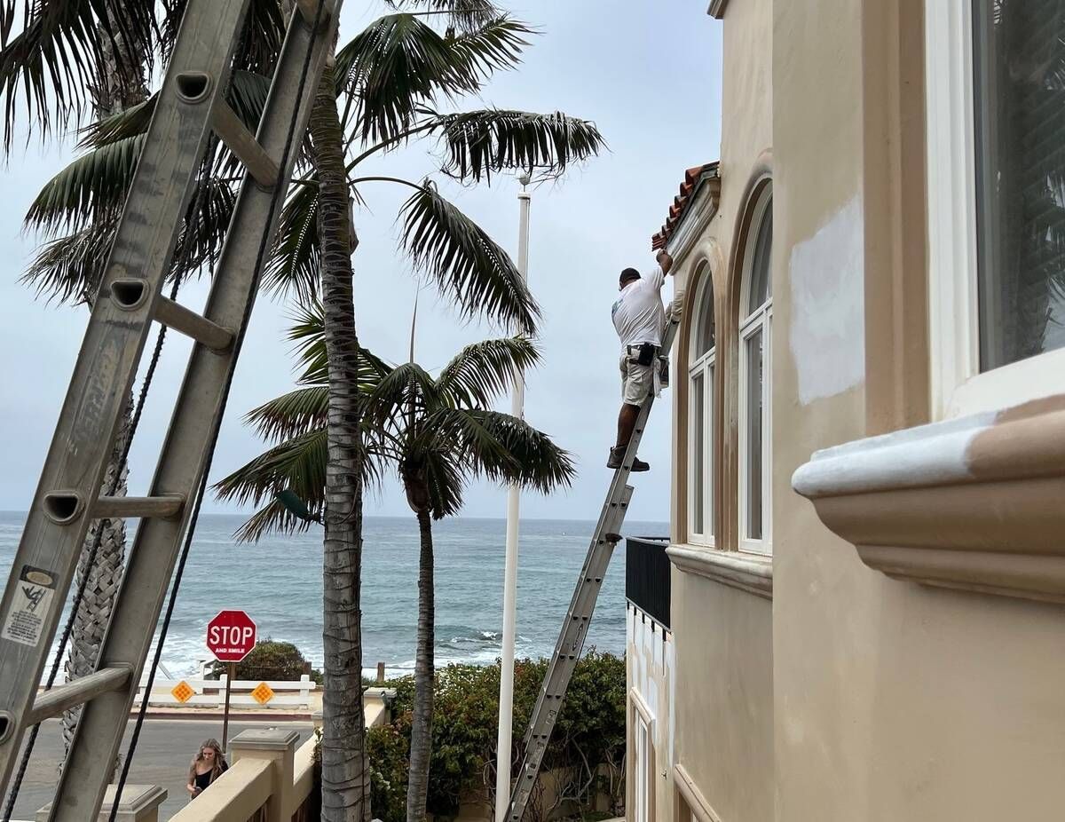 Person paints trim on building next to ocean. Two ladders lean against the building, stop sign and palm trees visible.