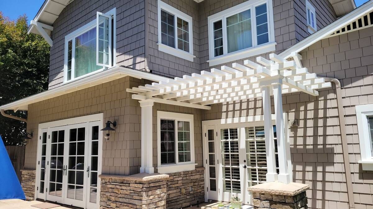 Two-story house with gray shingles, tan siding, white pergola, and French doors.