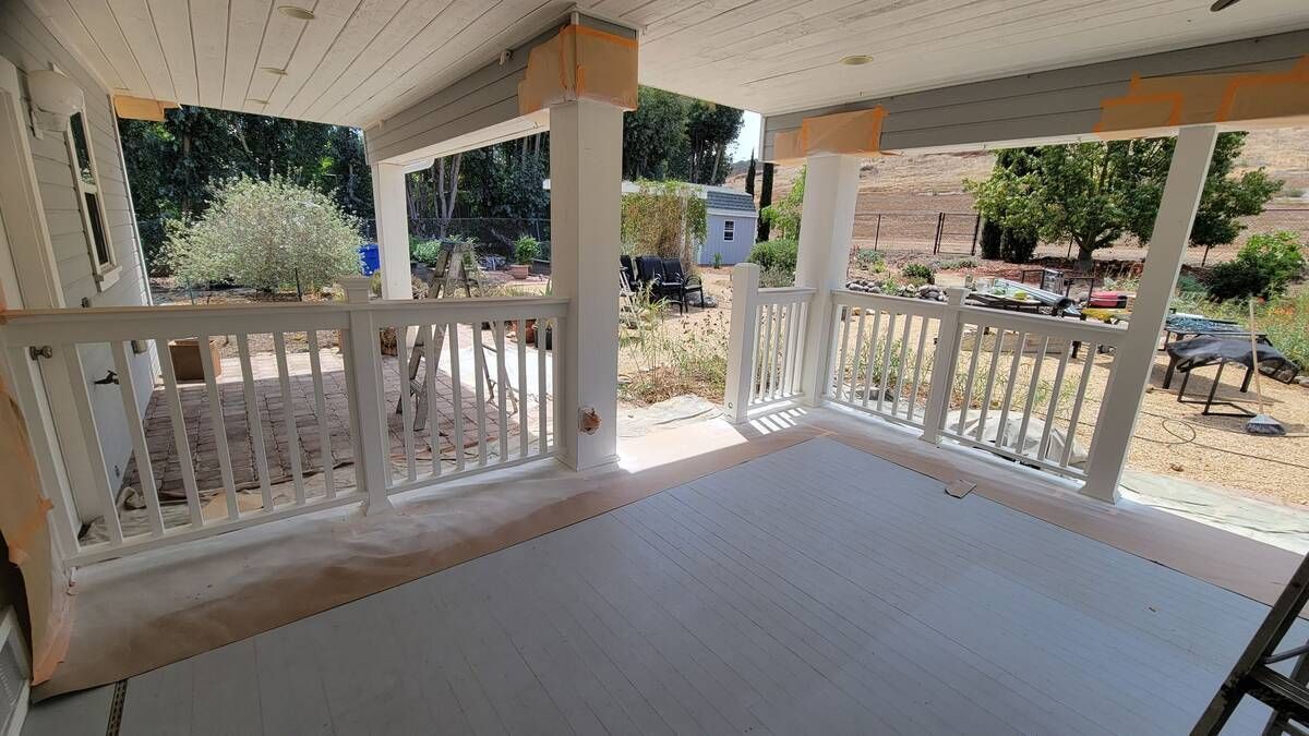 Covered porch with white railings and columns, overlooking a yard with trees and a small building.